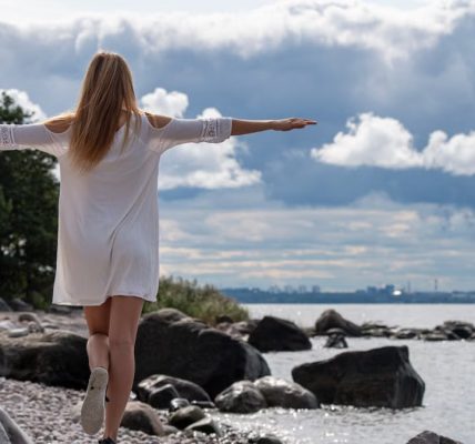 A woman in a white dress balances on rocks by the sea in Pringi, Estonia, under a cloudy sky.